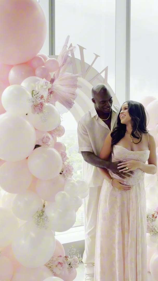 Expecting parents at a blush and white balloon arch under a floor-to-ceiling window