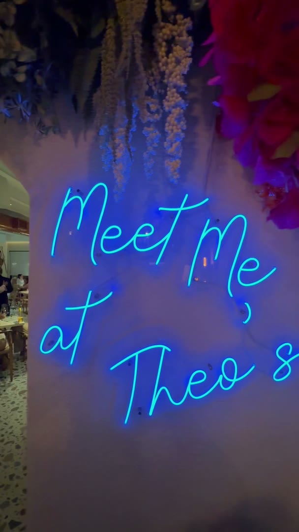 A neon Meet Me at Theo's sign under a hanging floral ceiling in a restaurant dining room