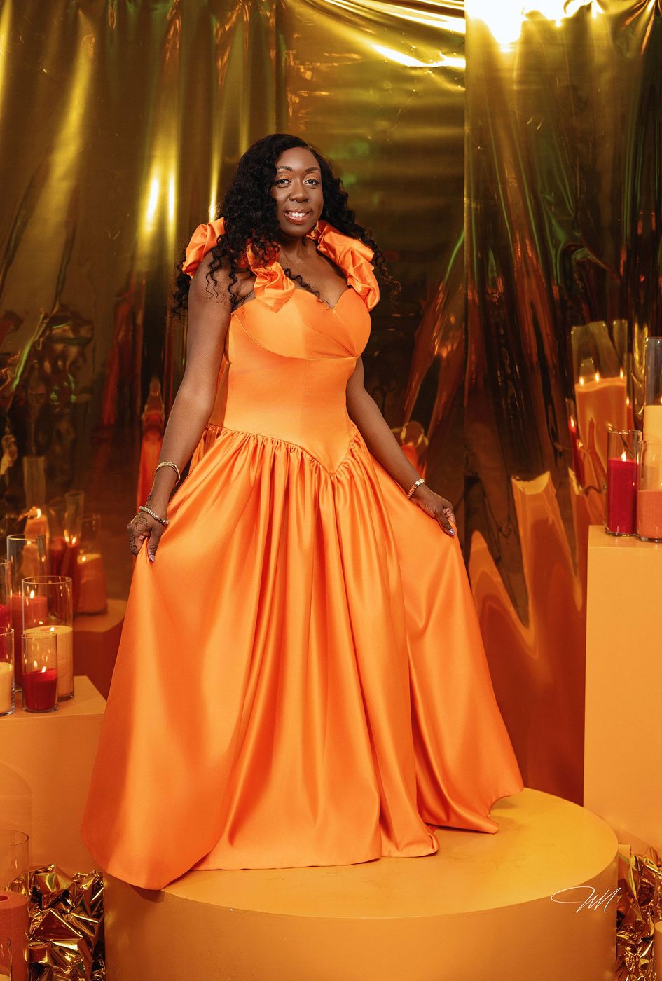 A guest in a full orange gown photographed against a gold foil backdrop