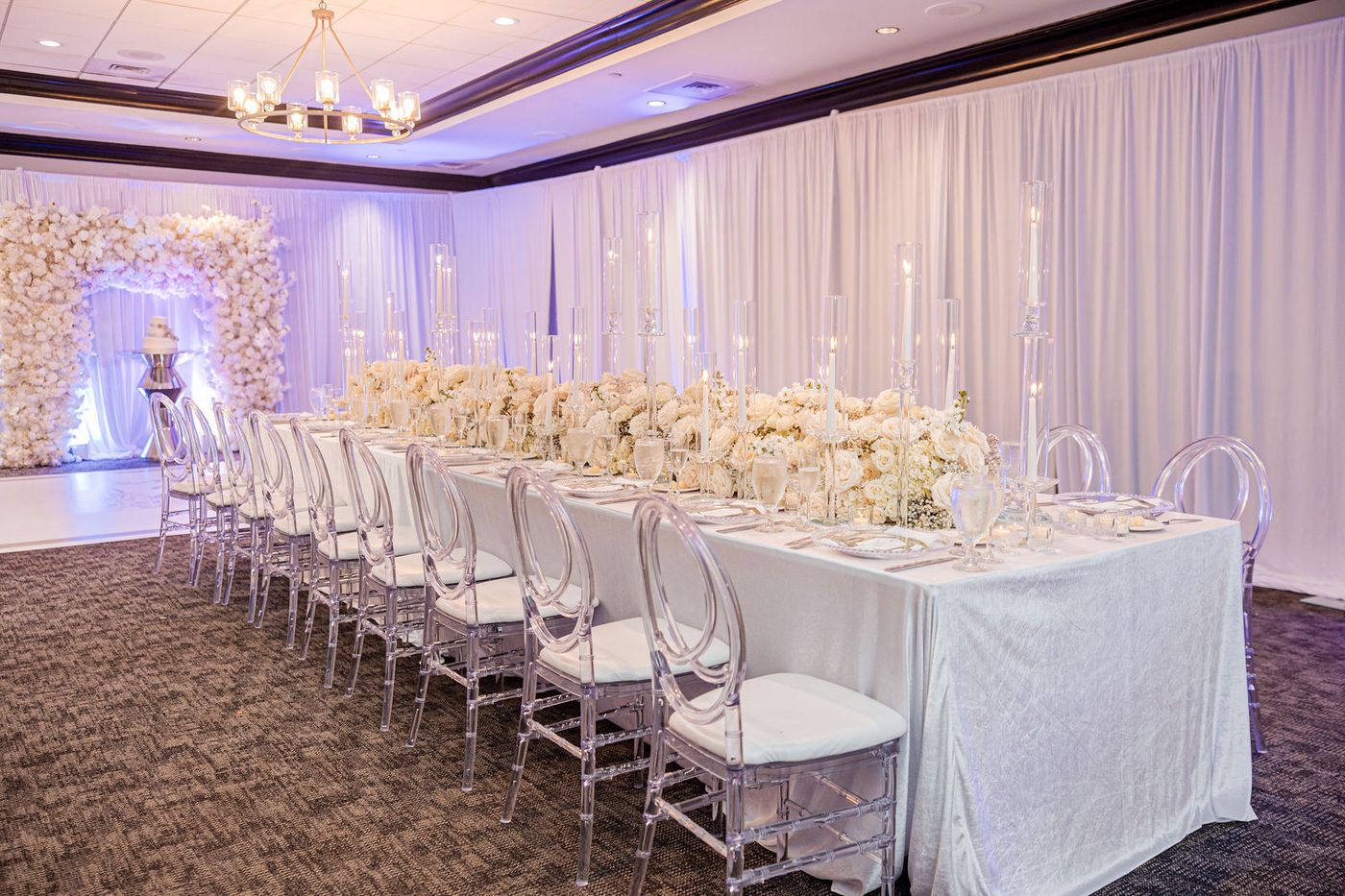 A long banquet table lined with white roses and tapered candles beneath a ballroom chandelier