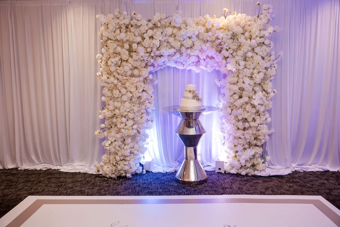 A white floral arch over a monogrammed dance floor, lit from behind in a hotel ballroom